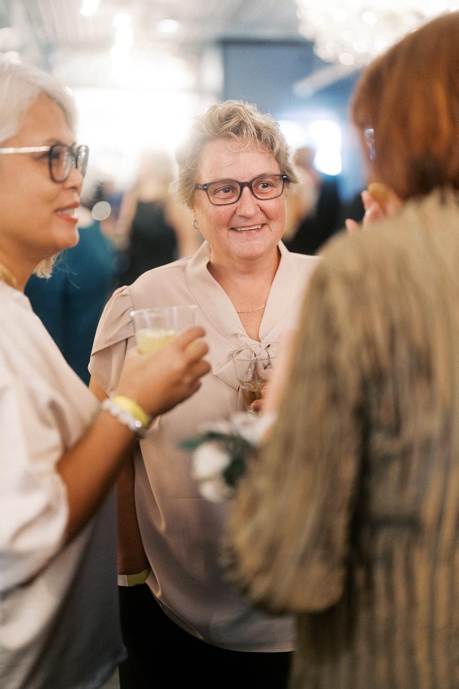 A group of women engaged in conversation at a social event, each holding a drink, with a bright, blurred background.