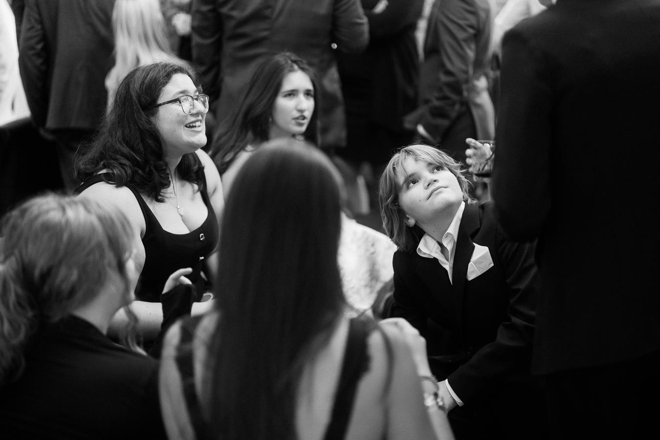 Black and white image of a social gathering, featuring a group of people dressed formally, engaged in conversation and laughter.