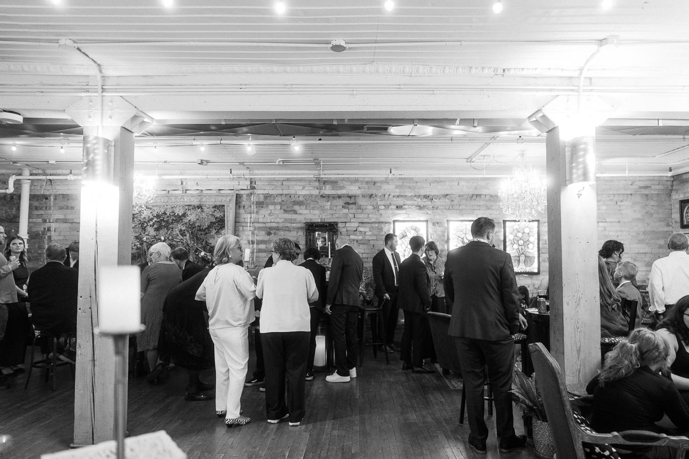 Black and white image of a social gathering in a rustic indoor venue with ambient lighting, featuring people mingling and conversing in formal attire.