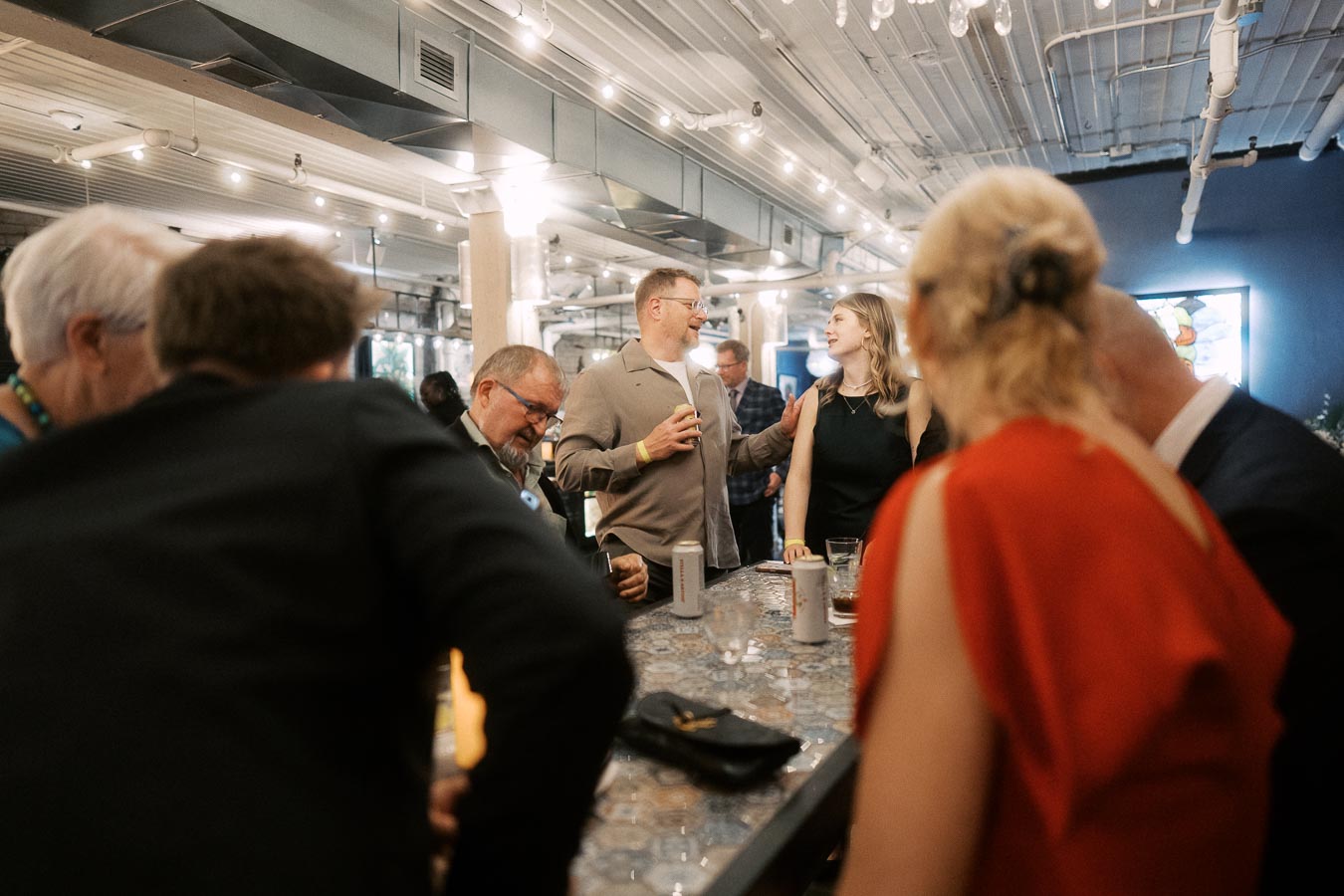 A group of people engaging in conversation at a modern indoor event or party, standing around a long table covered with drinks and personal items, under warm ambient lighting and stylish industrial decor.