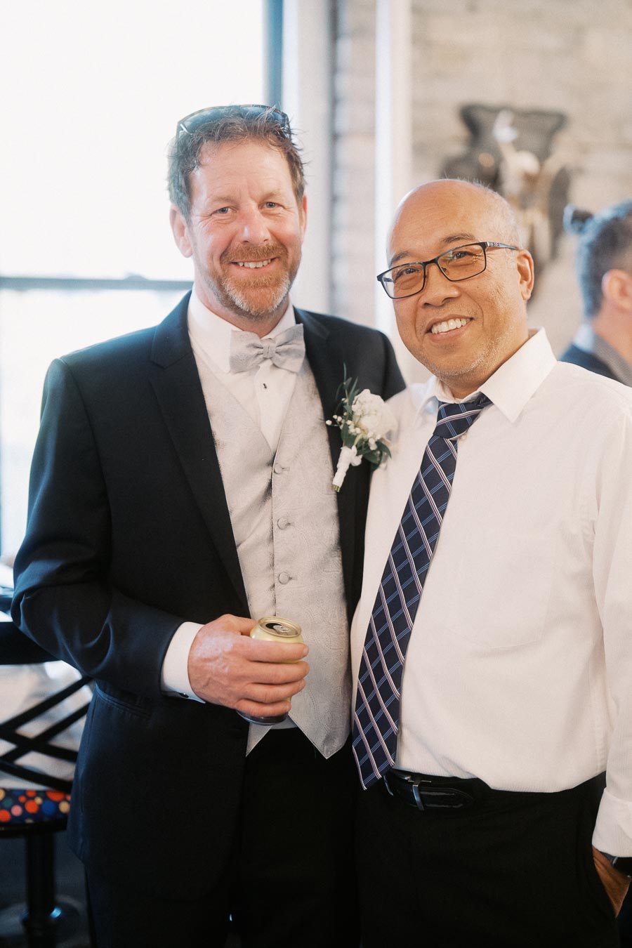Two smiling men posing together at a formal event, one wearing a suit with a boutonniere and holding a drink, the other in a shirt and tie, in an indoor setting with a window and decorative background.