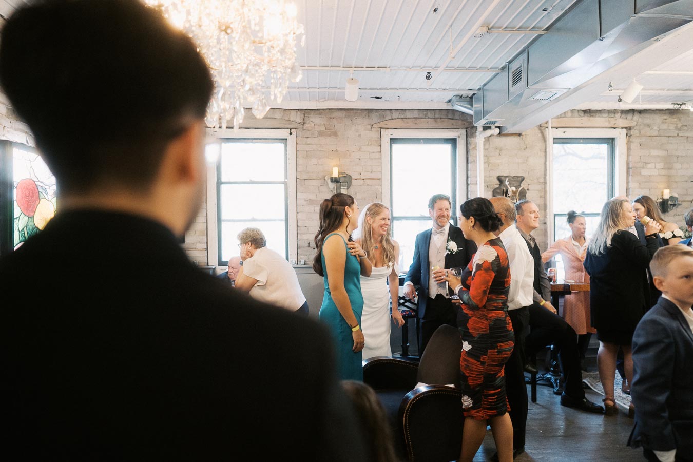 Wedding reception with guests socializing in a warmly lit venue. A bride in a white dress and a groom in a suit are seen smiling among friends. The room features exposed brick walls, large windows, and a decorative chandelier, creating a cozy and elegant atmosphere.