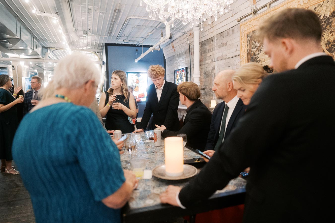 A diverse group of people enjoying a gathering in a stylish venue with modern decor and elegant lighting, featuring a prominently displayed chandelier and a variety of drinks on the table.