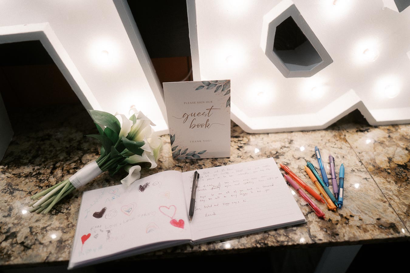 Wedding guest book display with open pages, colorful pens, and a bouquet of white flowers on a granite table, accompanied by illuminated letters in the background.