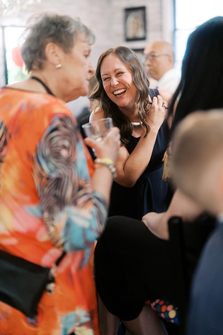 Smiling woman enjoying a social gathering, chatting with an older woman in a colorful dress, in a lively indoor setting.
