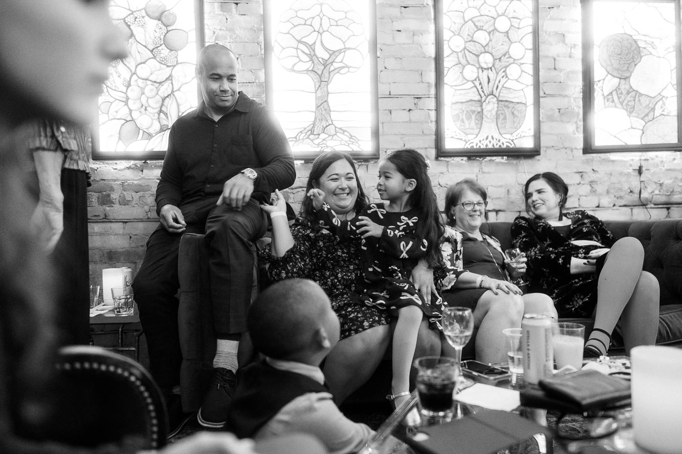 A black and white photo captures a lively gathering of people sitting on a couch in a cozy room with brick walls and decorative stained glass windows. The scene is filled with warmth and smiles as adults and children interact, highlighting a joyful and social atmosphere.