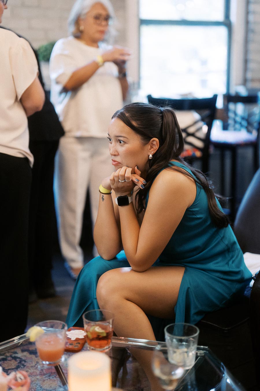 Woman in a turquoise dress sits at a social gathering, thoughtfully observing the room; background includes people mingling and a window, with cocktails on a glass table in the foreground.