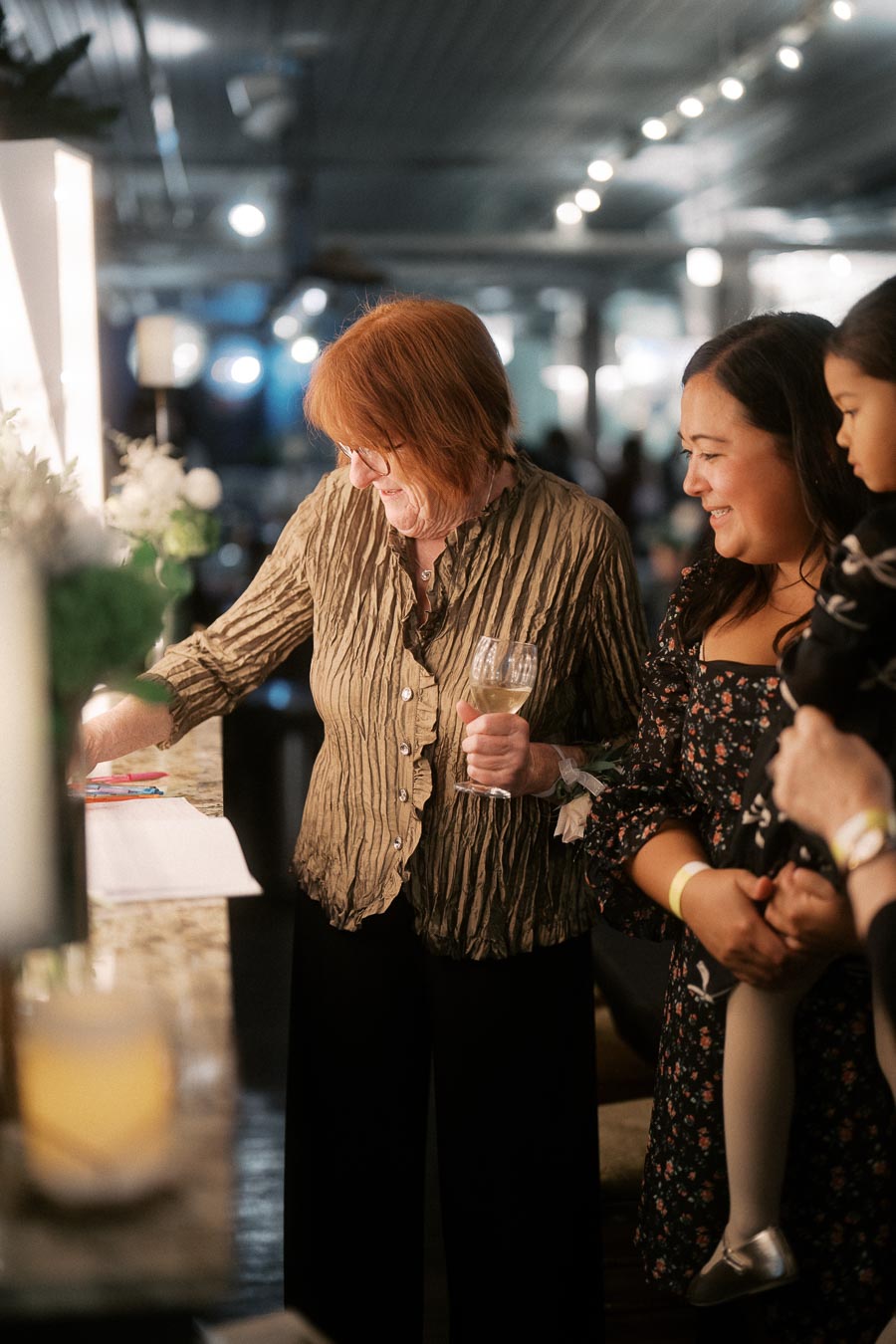 A smiling woman with red hair holding a glass of wine, standing next to a woman and child at a festive indoor gathering, illuminated by soft lighting in a lively setting.