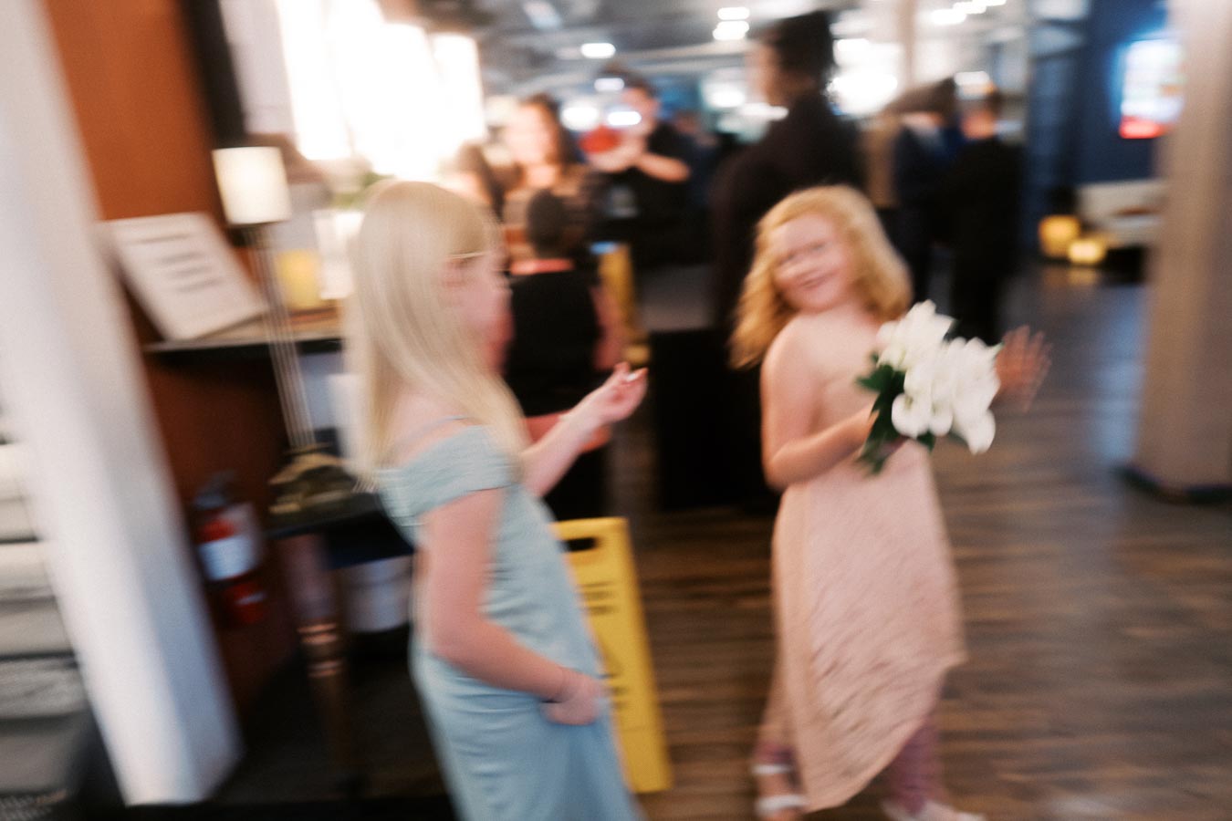 Blurry photo of two young girls in elegant dresses at a social event, one holding a bouquet of white flowers, with people and decorations in the background.