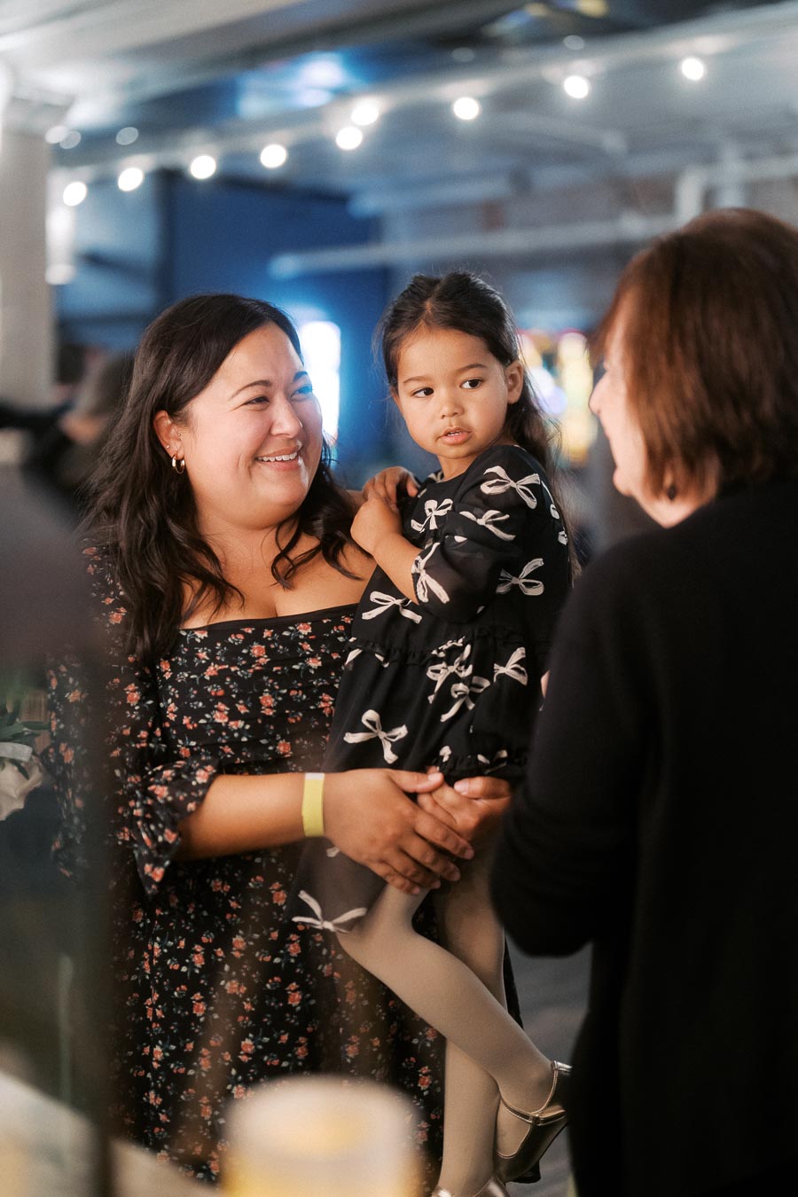 A smiling woman holding a young girl dressed in a cute bow-patterned dress, engaging in conversation with another person in an indoor setting with decorative lights.