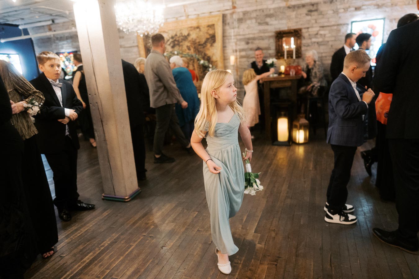 Young girl in a light blue dress holding a bouquet at a formal indoor gathering, surrounded by well-dressed guests in a warmly lit venue.