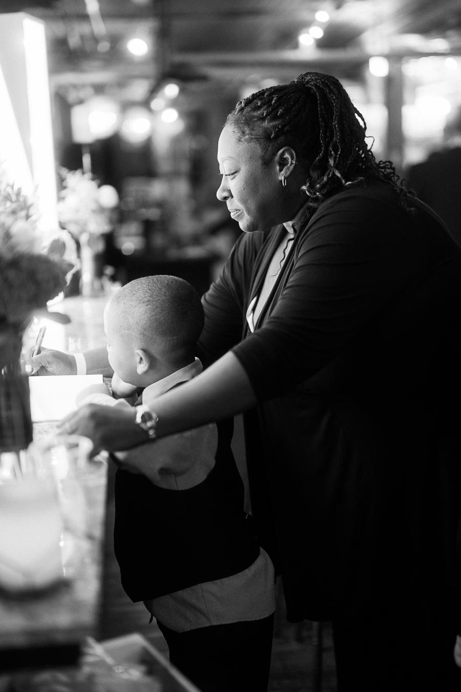 Black and white image of an adult helping a child write at a table, surrounded by blurred lights and flowers in the background.