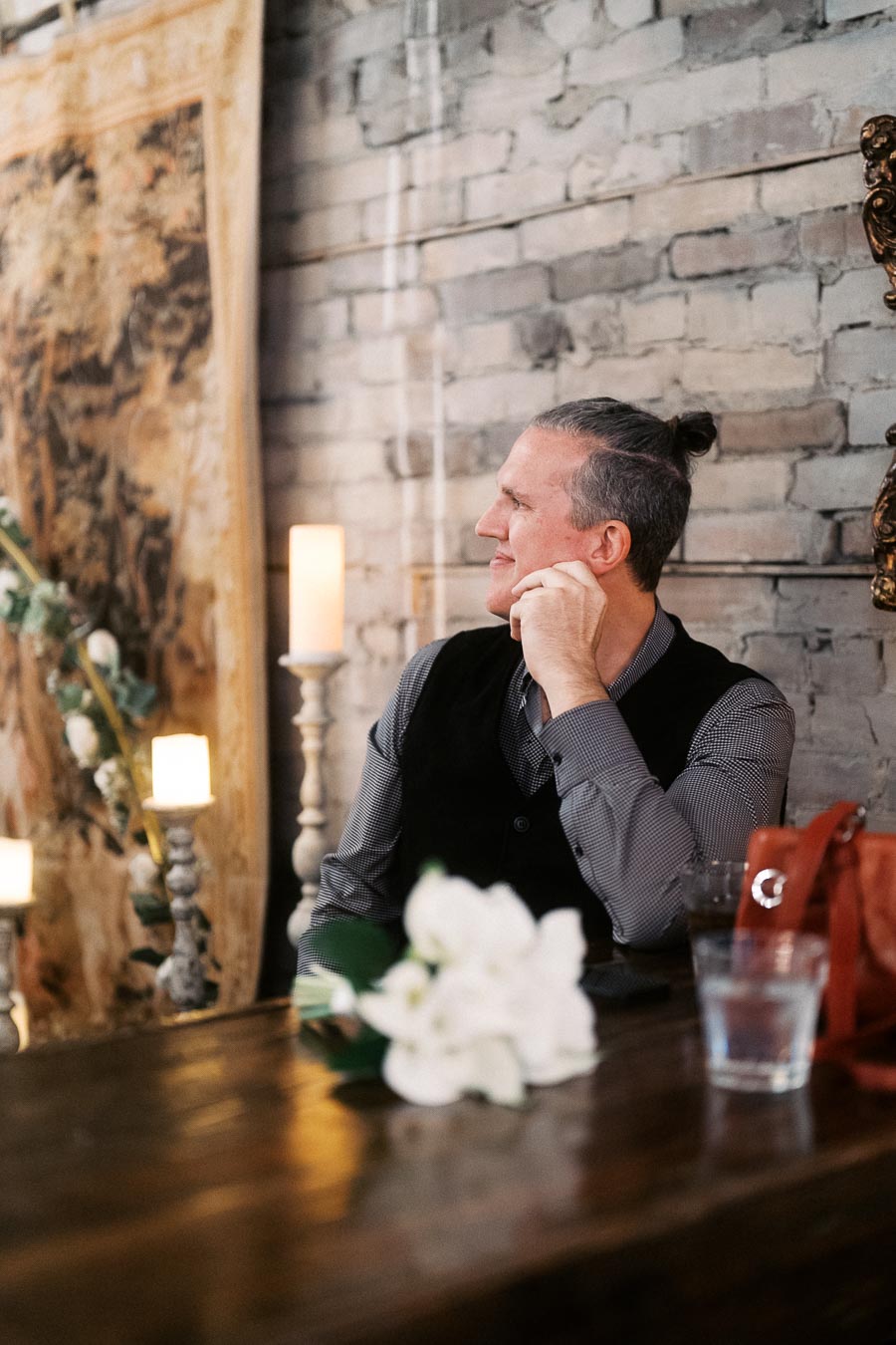 Man with a bun sitting at a rustic table, gazing thoughtfully to the side, surrounded by candles and a brick wall backdrop, with a bouquet of white flowers in the foreground.