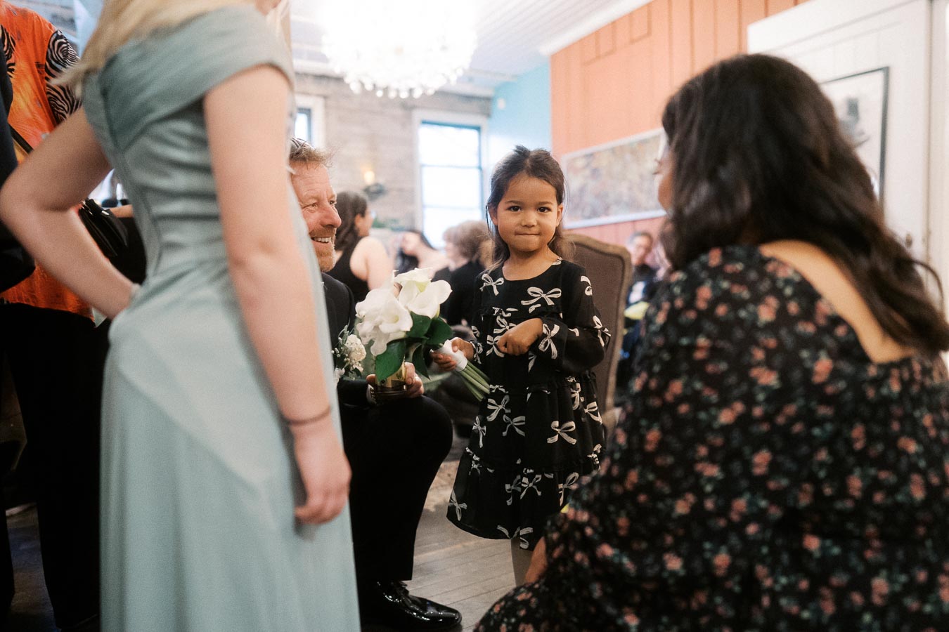 A young girl in a patterned dress stands in a warmly lit room, surrounded by a smiling man holding a bouquet of white flowers and elegantly dressed women. The setting suggests a joyful gathering or celebration.