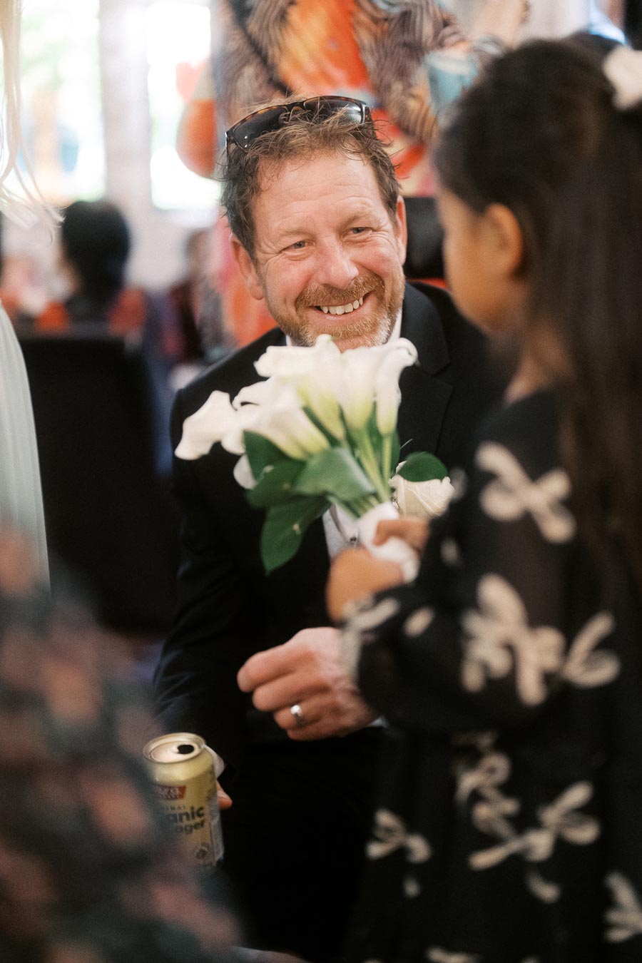 A man smiling warmly and talking to a child, who is holding a bouquet of white flowers, at a social gathering. He is wearing sunglasses on his head and holding a can in his left hand. The background is softly blurred with colorful artwork.