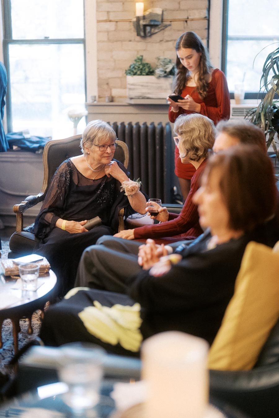 A group of people engaged in conversation at a cozy indoor gathering, with an elegantly dressed woman holding a drink, and a younger person in the background looking at a phone, set against a warmly lit living room backdrop.
