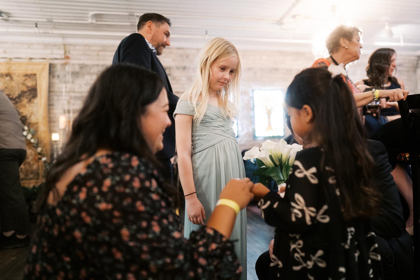 A group of people interacting at an indoor social gathering, with a young girl in a light blue dress holding a bouquet of white flowers, surrounded by adults and children in a warmly lit room.