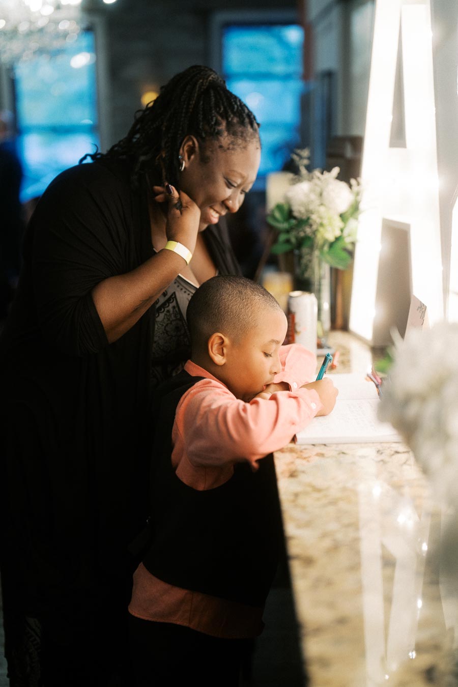 A woman smiling warmly as she watches a young boy intently writing at a countertop adorned with decorative elements and soft lighting.