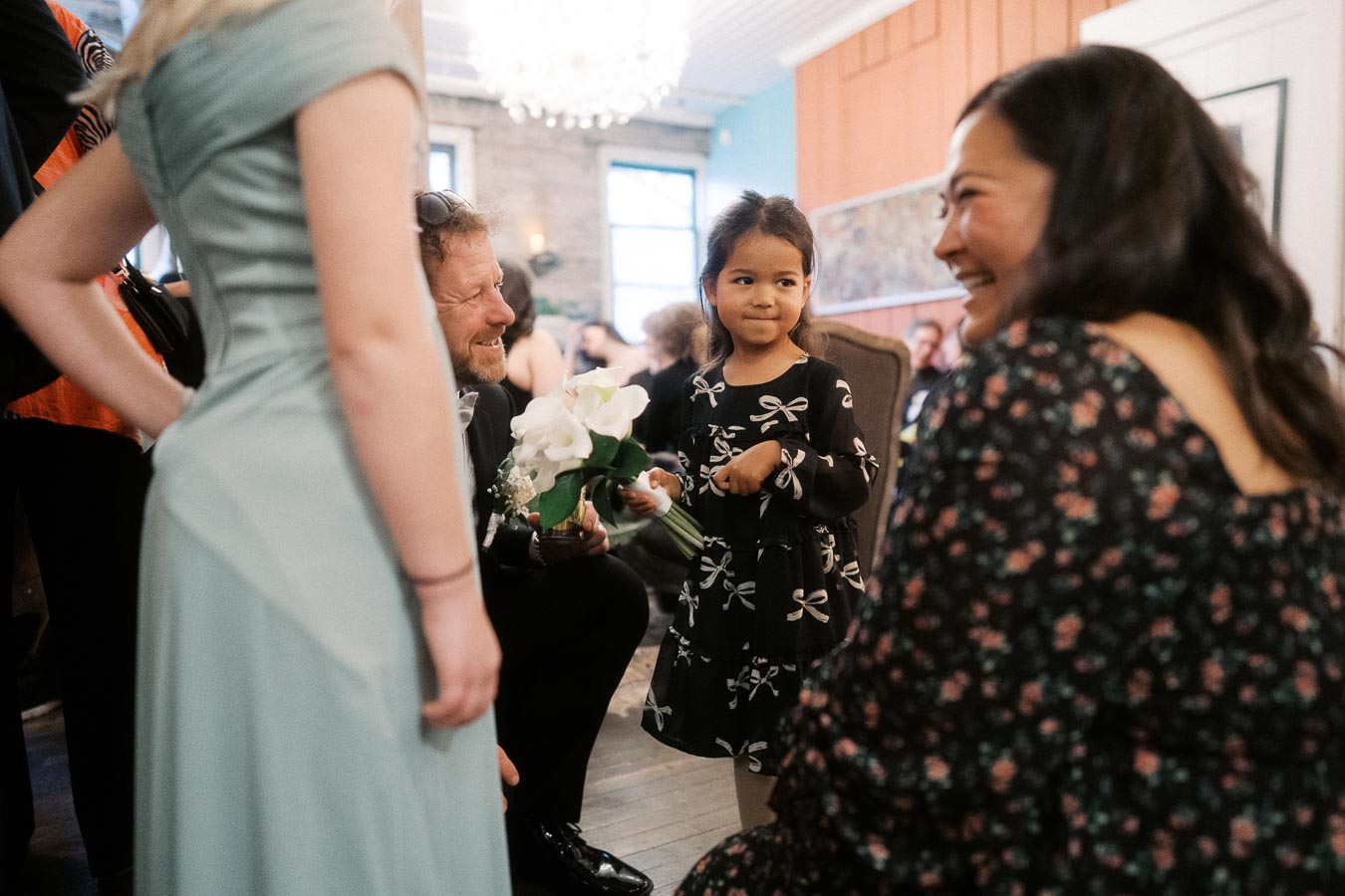 A joyful indoor gathering with a young girl in a navy dress holding a bouquet of flowers, surrounded by smiling adults in an elegantly decorated room.