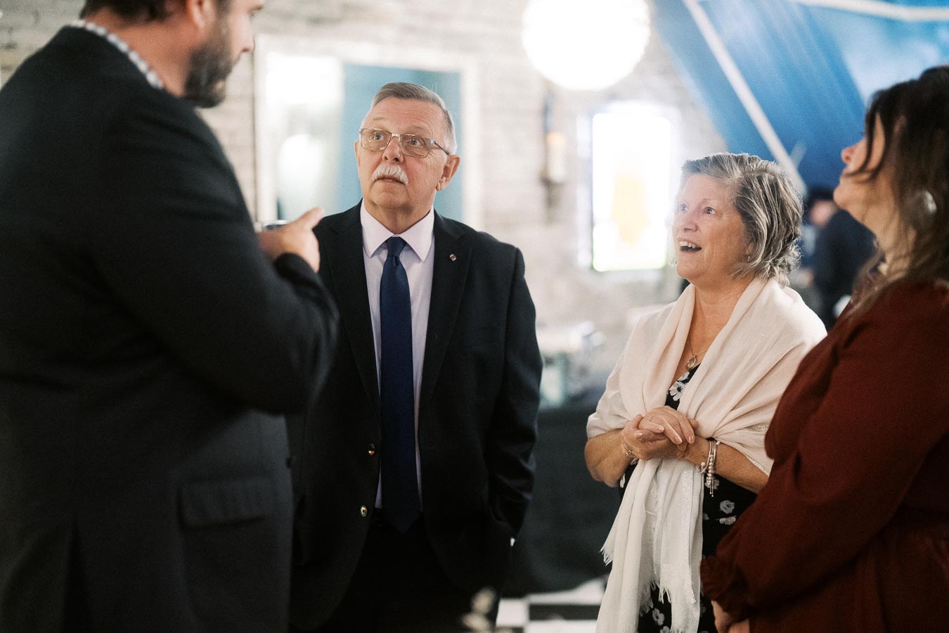 A group of people engaged in conversation at a formal event, featuring a man in a suit and tie, as well as a woman wearing a scarf, with a casual and friendly atmosphere.