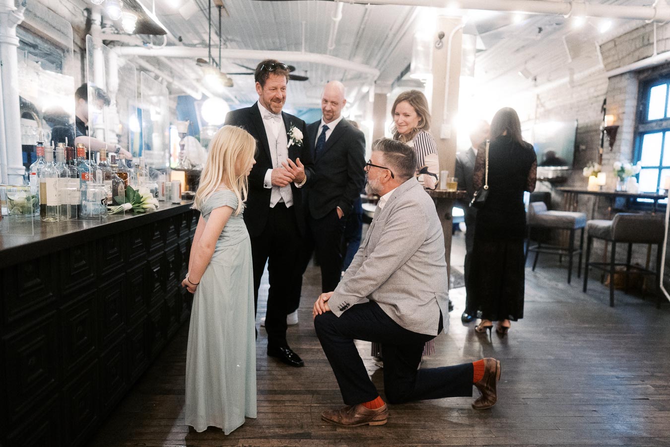 Group of people socializing at an elegant indoor event, with a kneeling man engaging with a young girl in a light blue dress, while others in formal attire stand nearby. The setting features a well-stocked bar and warm ambient lighting.