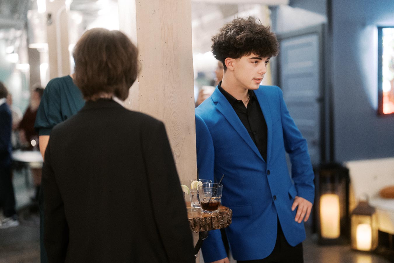 Young man in a vibrant blue suit leaning against a wooden pillar in a stylish indoor setting, engaged in conversation with another person in a black jacket. A round wooden shelf holds drinks with lime wedges in the foreground, suggesting a social gathering or event.