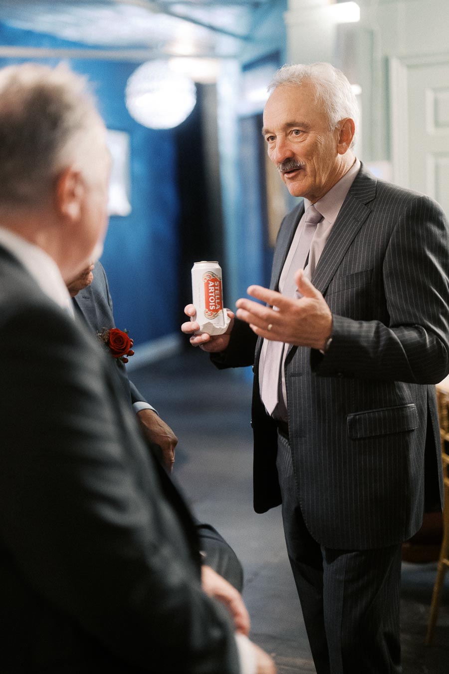 Man in a suit holding a Stella Artois can engaged in conversation at a social gathering.