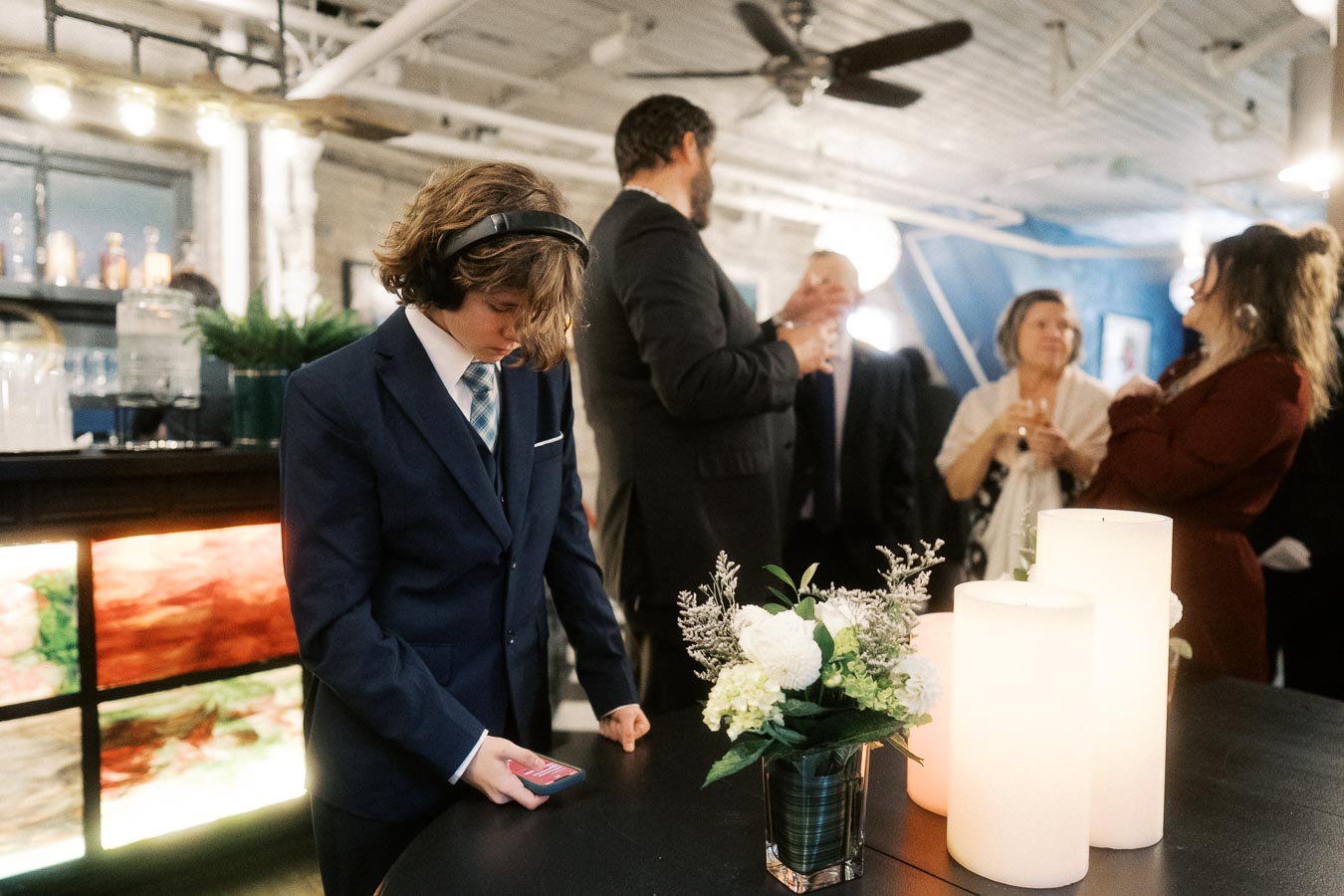 Young boy in a suit with headphones, looking at a phone during a social gathering in a modern event space, surrounded by adults conversing near decorative candles and flowers.