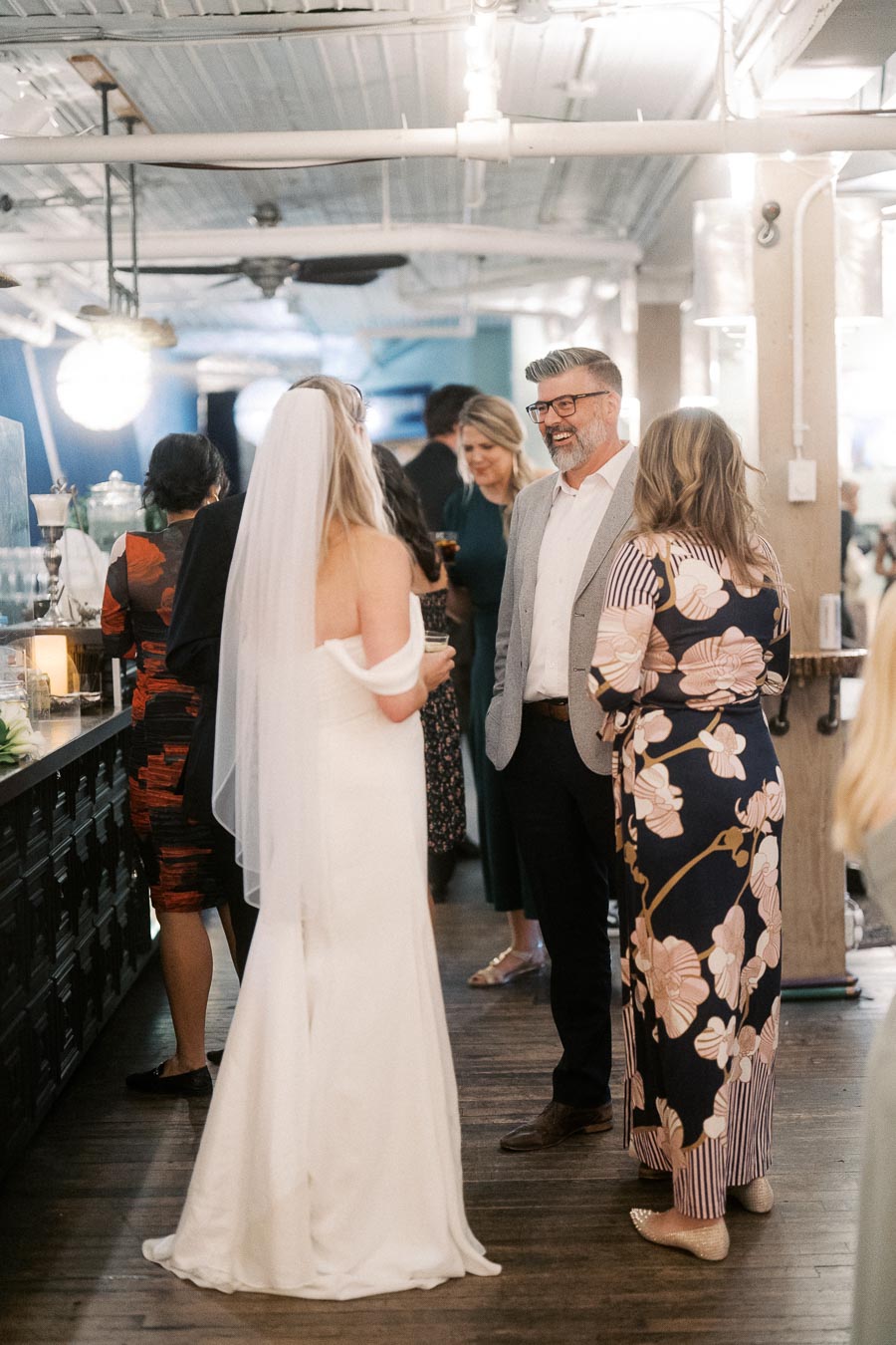 Wedding reception gathering with guests in elegant attire, including a bride in a white gown and veil, conversing in a warmly lit, stylish venue.