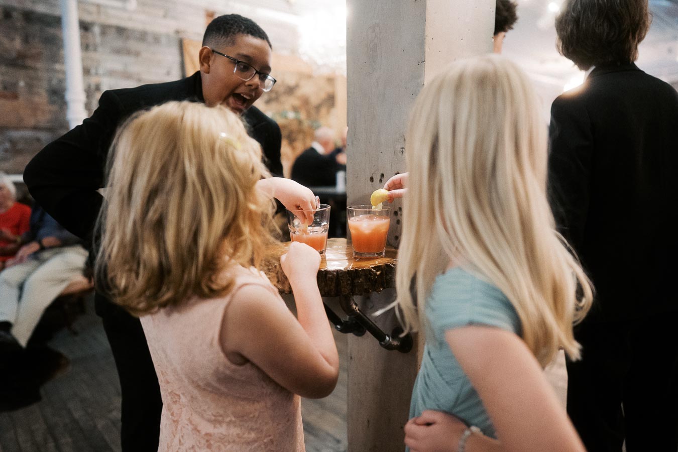 Children enjoying drinks at a rustic indoor event, with two girls and a boy interacting near a wooden table.