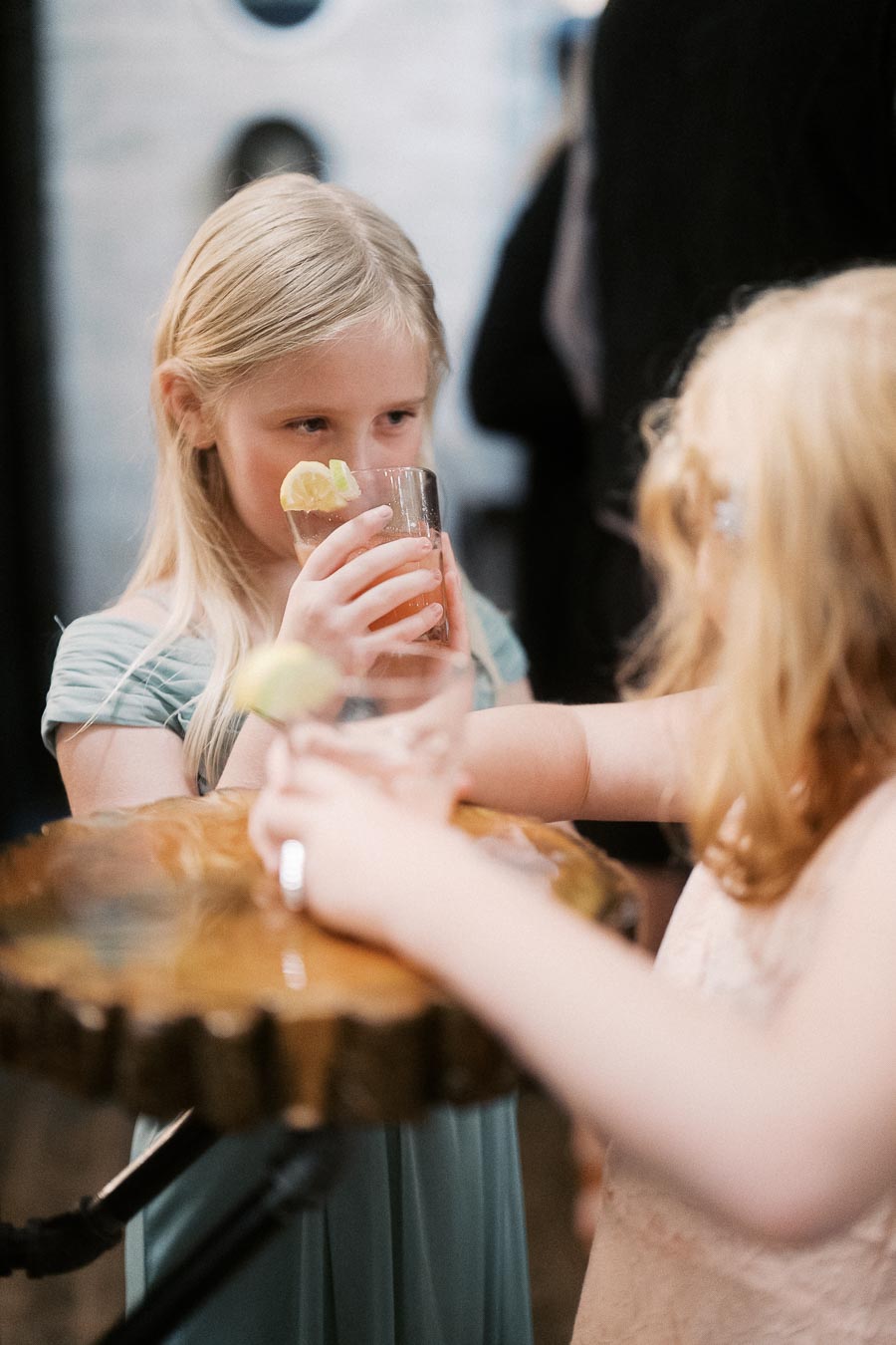 Two young girls enjoy drinks garnished with lemon at an indoor event, standing near a wooden table.