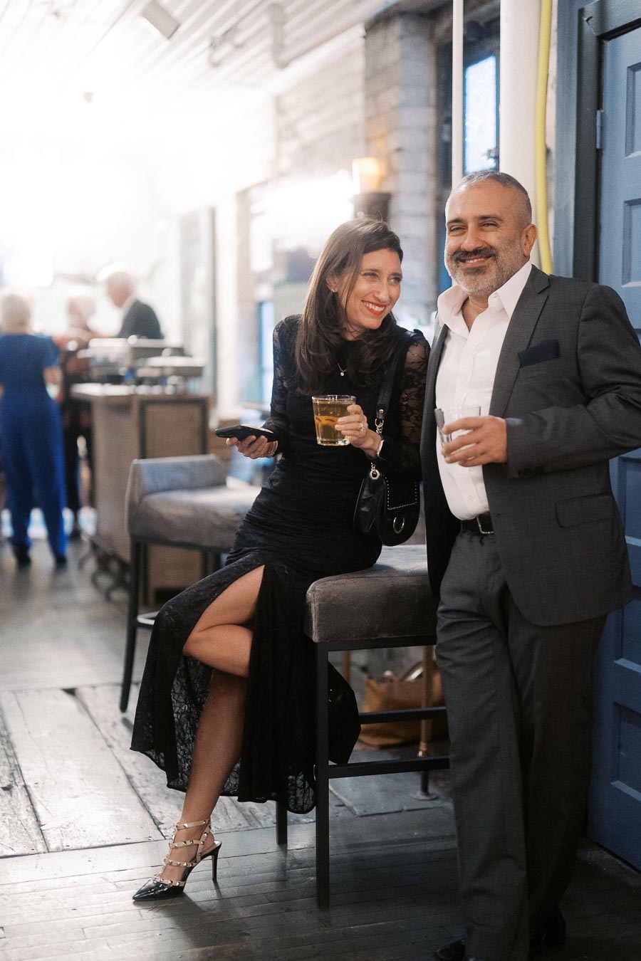 A woman in an elegant black dress and heels holds a drink and phone while smiling next to a man in a tailored suit, both enjoying a lively evening at a bar or lounge.