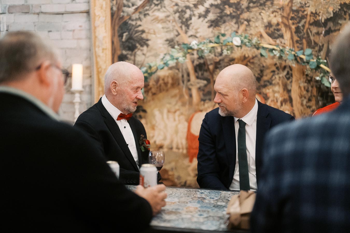 Two men in formal attire seated at a table, engaged in conversation during an indoor event, with a decorative tapestry in the background.