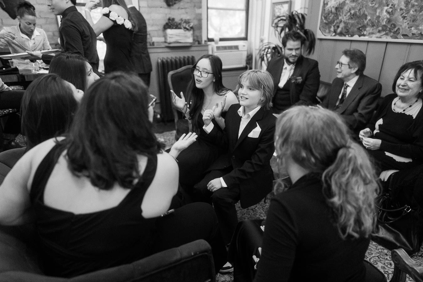 A black and white photo of people gathered at a social event, engaging in lively conversation. The group includes men, women, and children dressed in formal attire, seated in a warmly decorated room with artwork and plants.