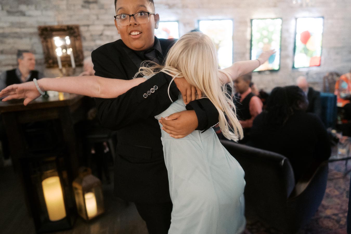 A joyful moment captured at a social gathering, featuring a boy in a black suit playfully hugging a girl in a light blue dress, with blurred guests and decorative lanterns in the background.