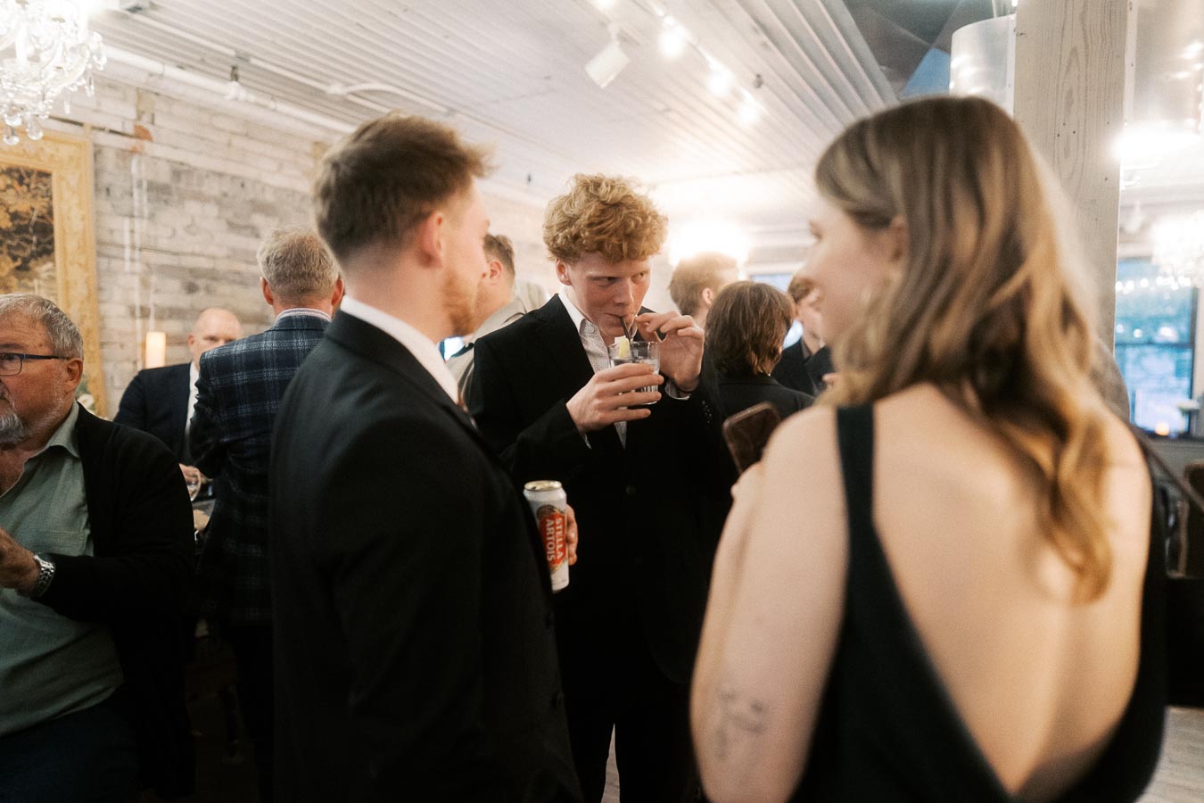 Group of people socializing at a sophisticated indoor event, with drinks in hand, dressed in formal attire, in a warmly lit venue with exposed brick walls and chandeliers.