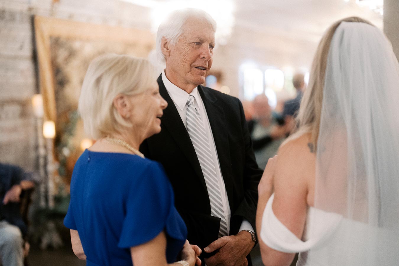 Elderly couple smiling and talking to a bride at a wedding ceremony, with elegant indoor setting and soft lighting.