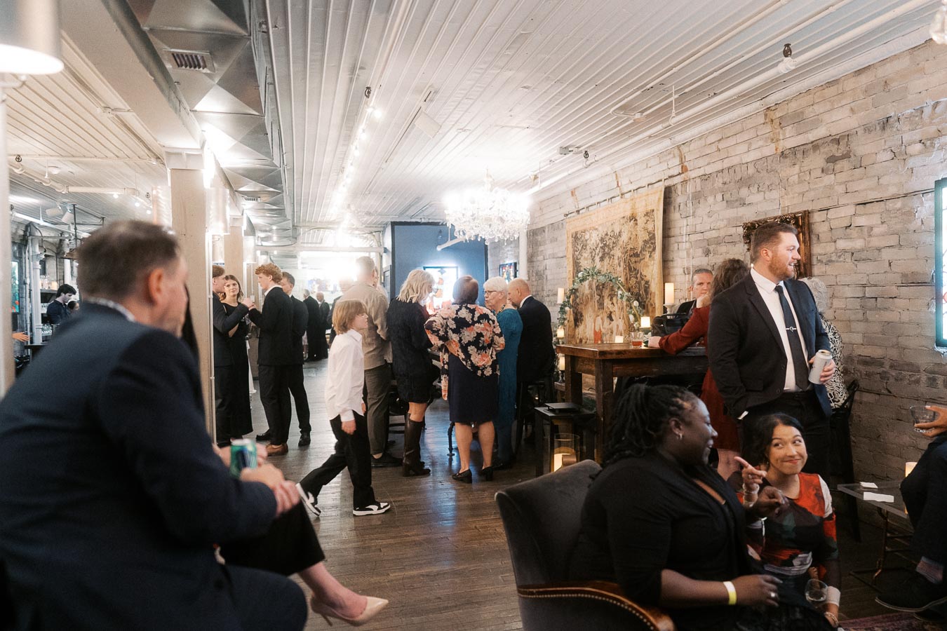 Gathering of people in formal attire socializing at an indoor event venue, featuring elegant decor with chandeliers and exposed brick walls. Guests are mingling and enjoying beverages, creating a lively and sophisticated atmosphere.