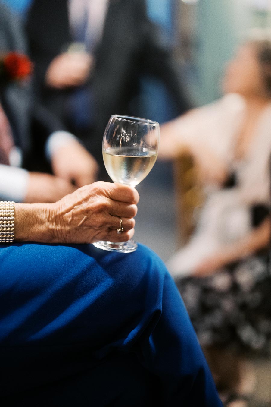 Elderly person holding a glass of white wine at a social gathering, wearing a pearl bracelet and blue clothing, with blurred people in the background.