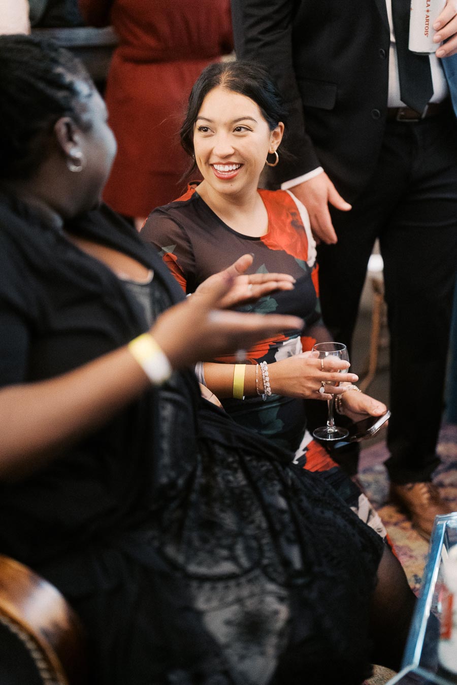Two people engaged in lively conversation at a social event, both smiling warmly. The scene is vibrant with one holding a wine glass and another gesturing expressively, suggesting a friendly and joyful atmosphere.