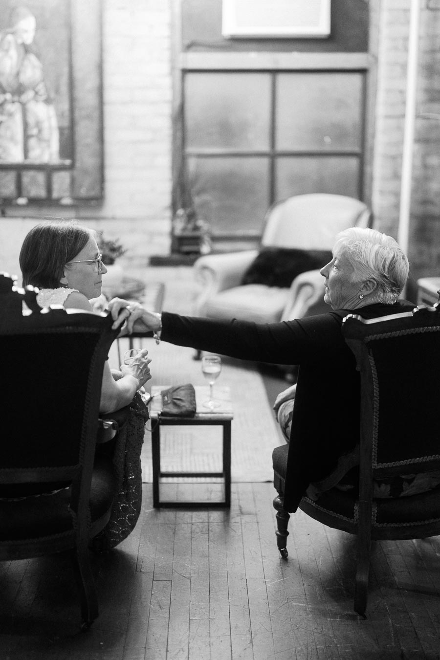 Black and white image of two elderly women sitting in a cozy living room, engaging in conversation with glasses of wine on a small table.