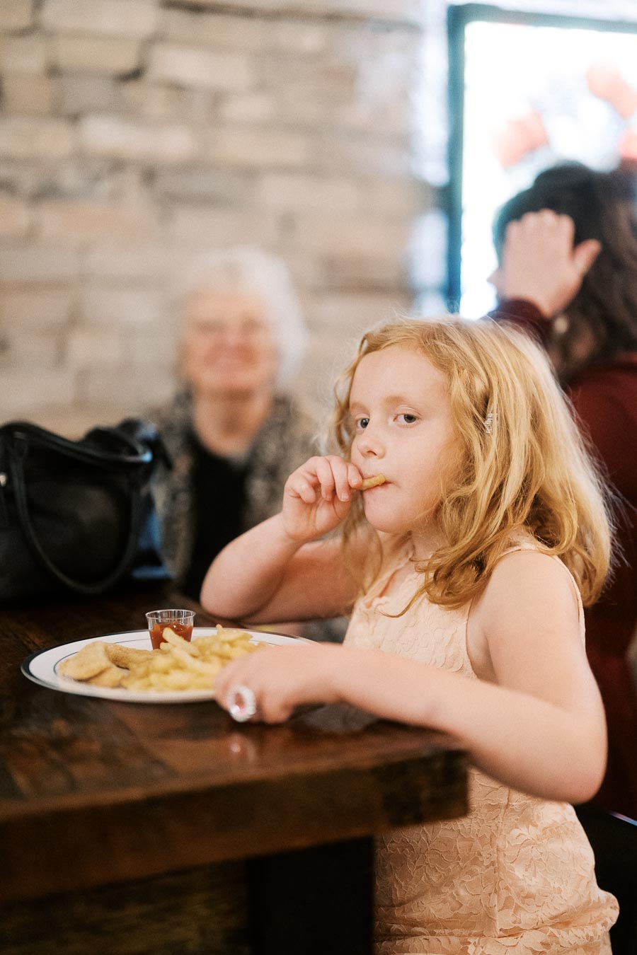 Young girl enjoying a meal of chicken nuggets and fries at a restaurant, with family in the background.