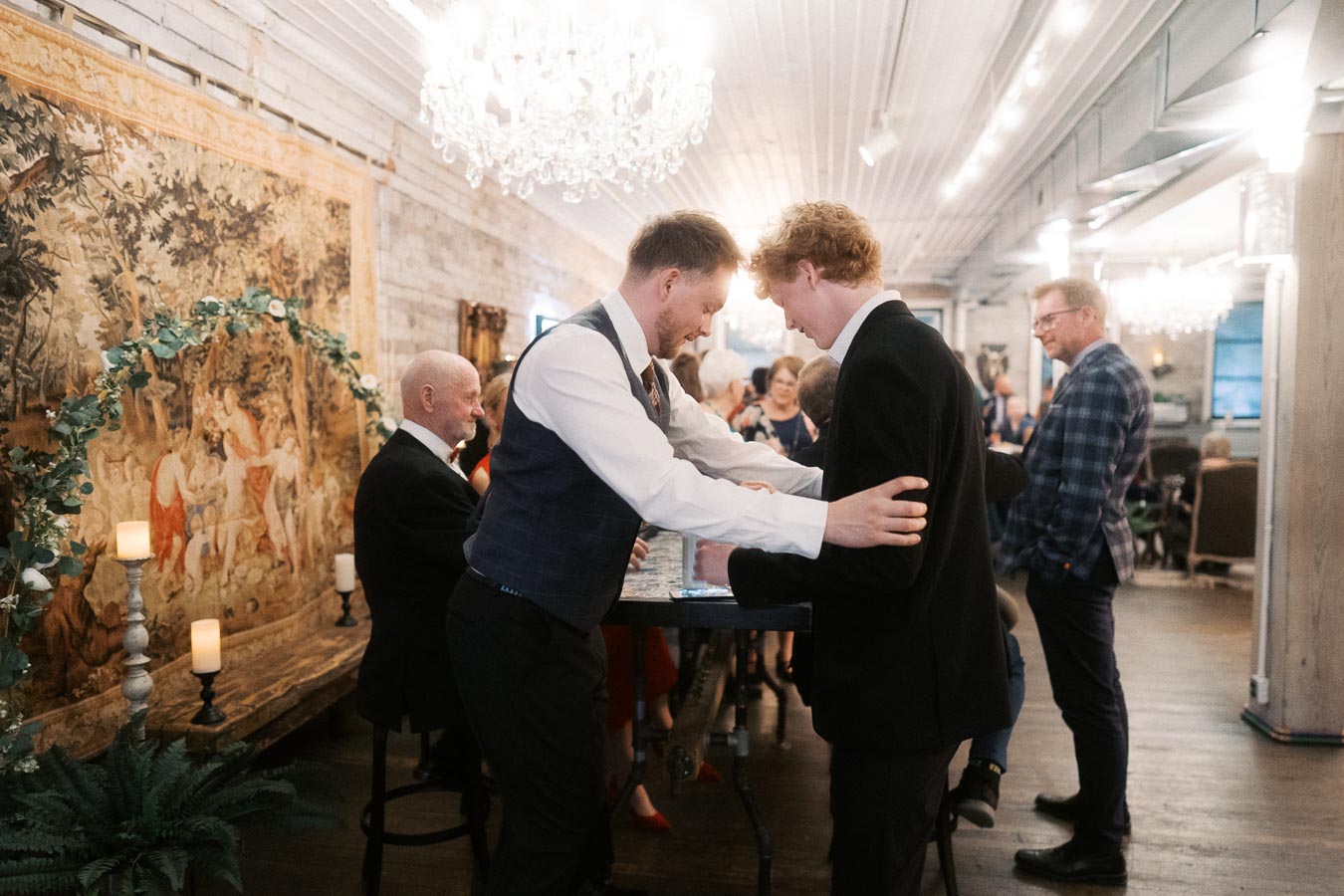 Group of people mingling at a formal event in an elegant room with chandeliers and a decorative tapestry, featuring two men engaged in a warm handshake in the foreground.