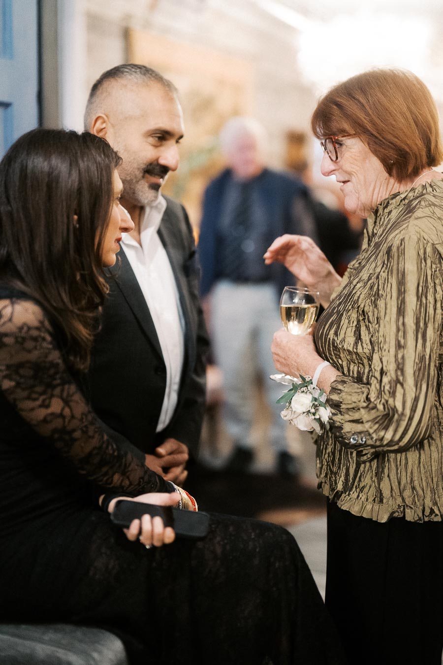 Elegant social gathering with three people engaged in conversation, featuring a woman in a stylish lace dress, a man in a formal suit, and an older woman wearing a fashionable blouse, holding a glass of white wine.