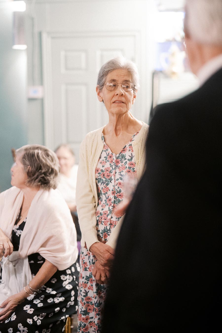 Elderly woman in floral dress and cardigan attentively engaging at a social gathering indoors, with people in the background.