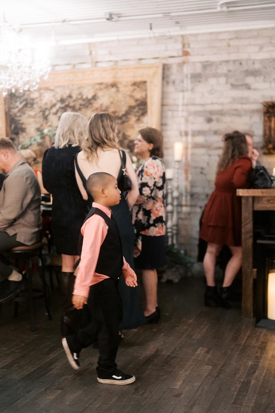 A young boy in a pink shirt and black vest walks through a social gathering in a stylish venue, where adults are engaged in conversation and enjoying the ambiance.