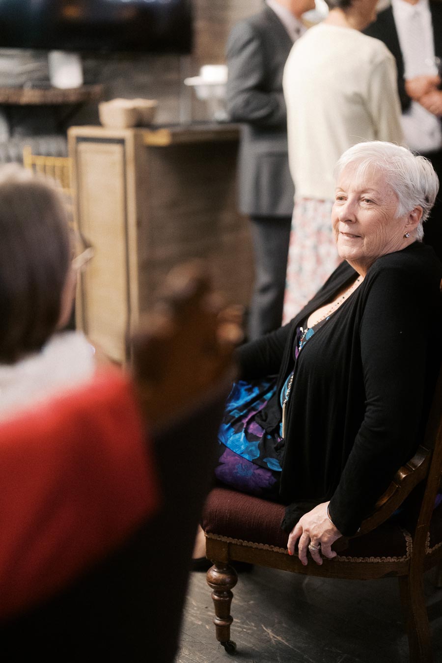 Elderly woman sitting on a chair, engaged in a social gathering with others in a warmly lit room.