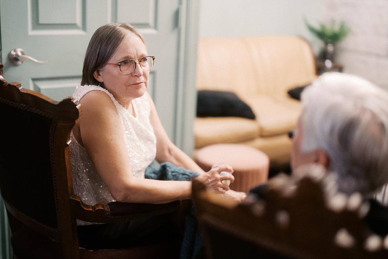 Elderly woman with glasses sitting in a chair having a conversation in a cozy room with a beige couch and soft lighting.