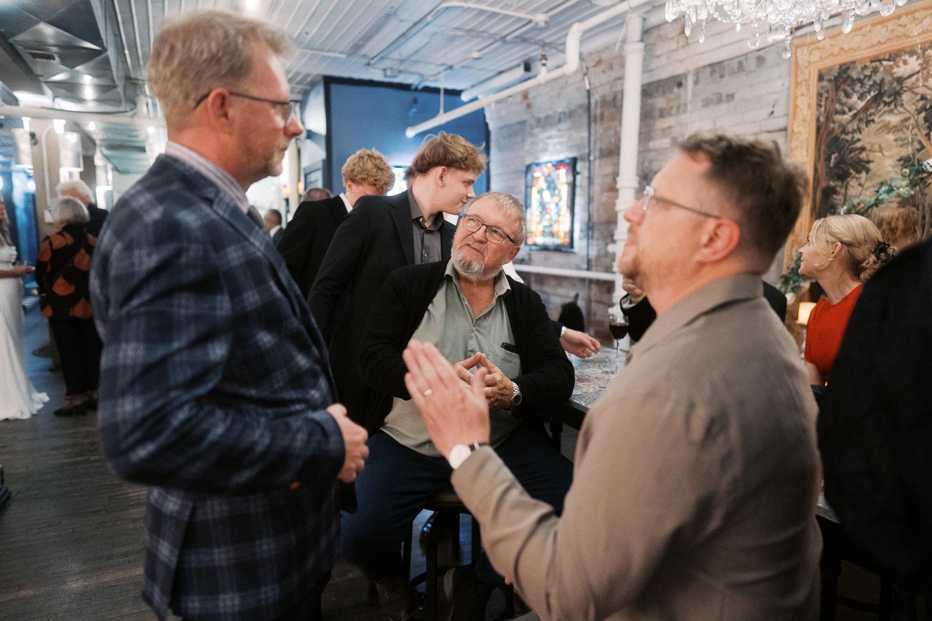 A group of people engaged in conversation at a social event inside a stylish venue, with a mix of casual and formal attire, visible wall art, and ambient lighting.
