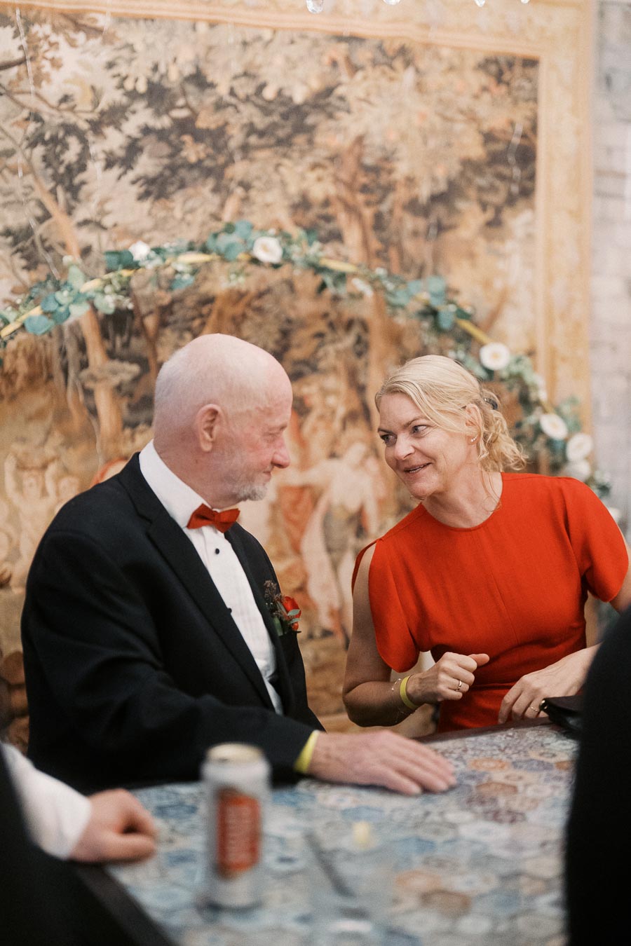 An elegantly dressed couple smiles warmly while seated at a mosaic-tiled table, set against a backdrop of a tapestry depicting a forest scene.